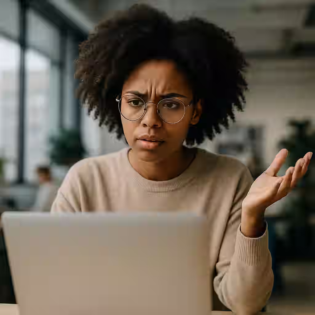 A woman wearing glasses and a beige sweater looks frustrated while sitting in front of a laptop, raising one hand in confusion or disbelief in a modern office environment.